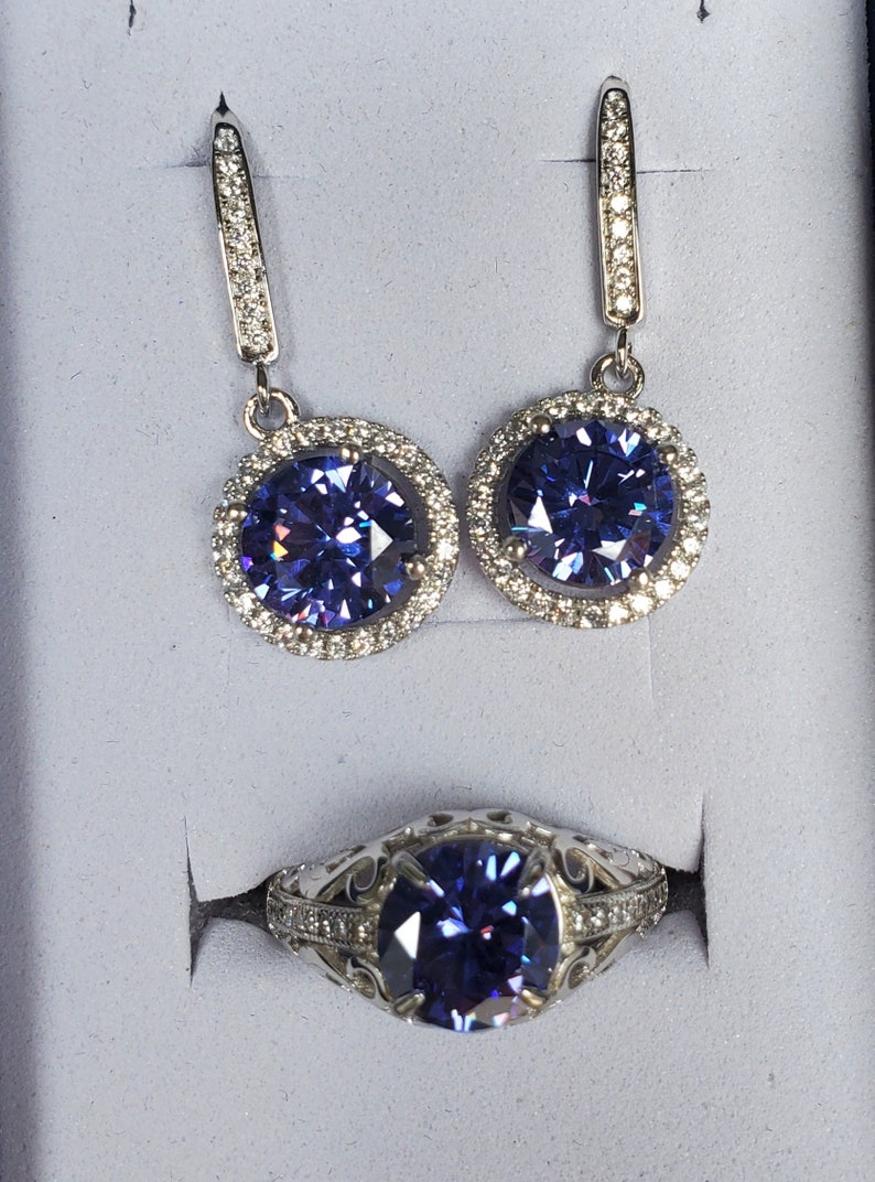 May include: A set of silver earrings and a ring featuring a large, round, purple gemstone encircled by smaller, clear gemstones.