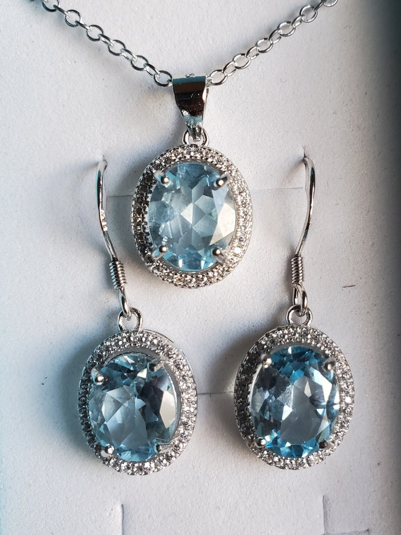 May include: A jewelry set featuring a silver necklace with a pendant and matching earrings. The pendant and earrings have oval, light blue gemstones surrounded by a halo of small, clear stones. The set is displayed on a white surface.