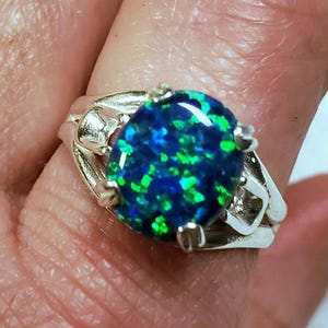 May include: A silver ring with a large, oval-shaped blue and green opal gemstone. The ring has a delicate, intricate design.