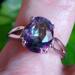 May include: A silver-toned ring featuring a large, oval-cut, multi-colored gemstone with iridescent flashes of purple, blue, and green. The stone is held in a four-prong setting, and the band has a split design on each side of the central stone.