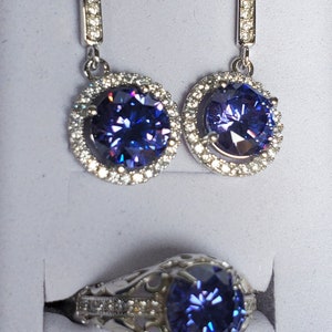 May include: A set of silver earrings and a ring featuring a large, round, purple gemstone encircled by smaller, clear gemstones.