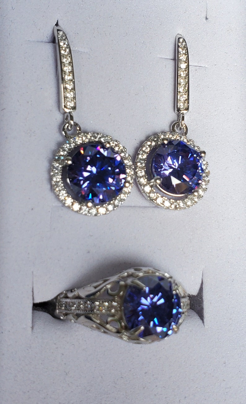 May include: A set of silver earrings and a ring featuring a large, round, purple gemstone encircled by smaller, clear gemstones.