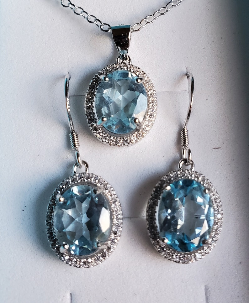May include: A jewelry set featuring a pendant necklace and matching earrings. The set includes an oval, light blue gemstone pendant and earrings, each surrounded by a halo of small, clear stones. The jewelry is set in a silver-colored metal.