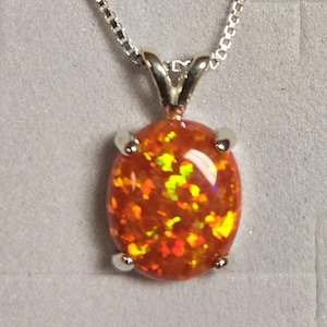 May include: An oval-shaped orange fire opal pendant set in a four-prong silver metal mounting, suspended from a delicate silver box chain necklace. The gemstone displays vibrant flashes of yellow and red light across its polished surface.