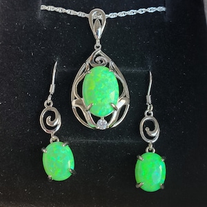 May include: A silver necklace and earrings set with green opal stones. The necklace features a teardrop-shaped pendant with a delicate floral design. The earrings are simple and elegant, with a small green opal stone hanging from a silver hook.