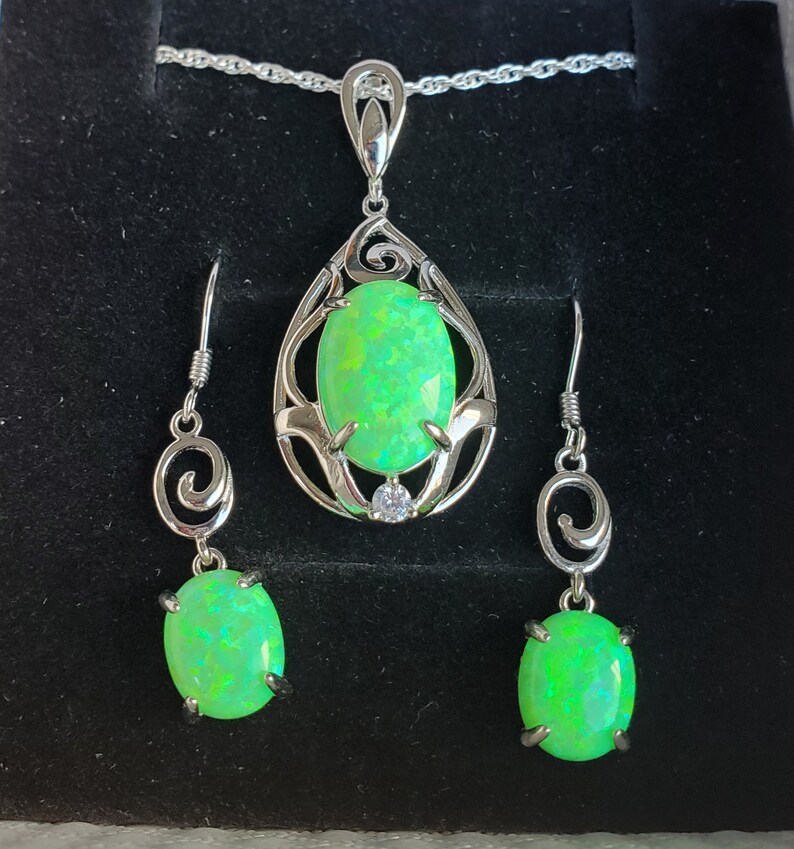 May include: A silver necklace and earrings set with green opal stones. The necklace features a teardrop-shaped pendant with a delicate floral design. The earrings are simple and elegant, with a small green opal stone hanging from a silver hook.