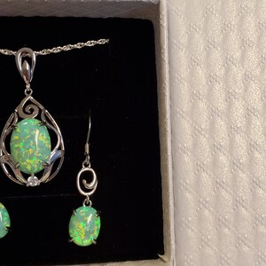 May include: A silver necklace and earrings set with a green opal pendant and matching earrings. The pendant and earrings feature a delicate, swirling design.