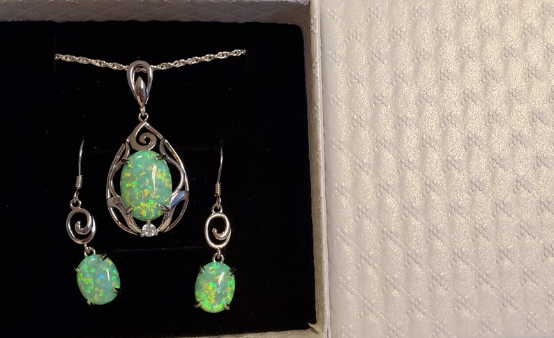 May include: A silver necklace and earrings set with a green opal pendant and matching earrings. The pendant and earrings feature a delicate, swirling design.