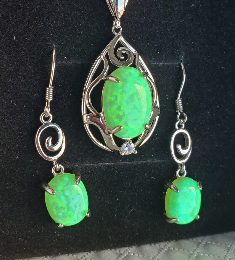May include: A silver necklace and earrings set with green opal stones. The necklace features a teardrop-shaped pendant with a delicate, swirling design. The earrings are simple and dangle with a small opal stone.
