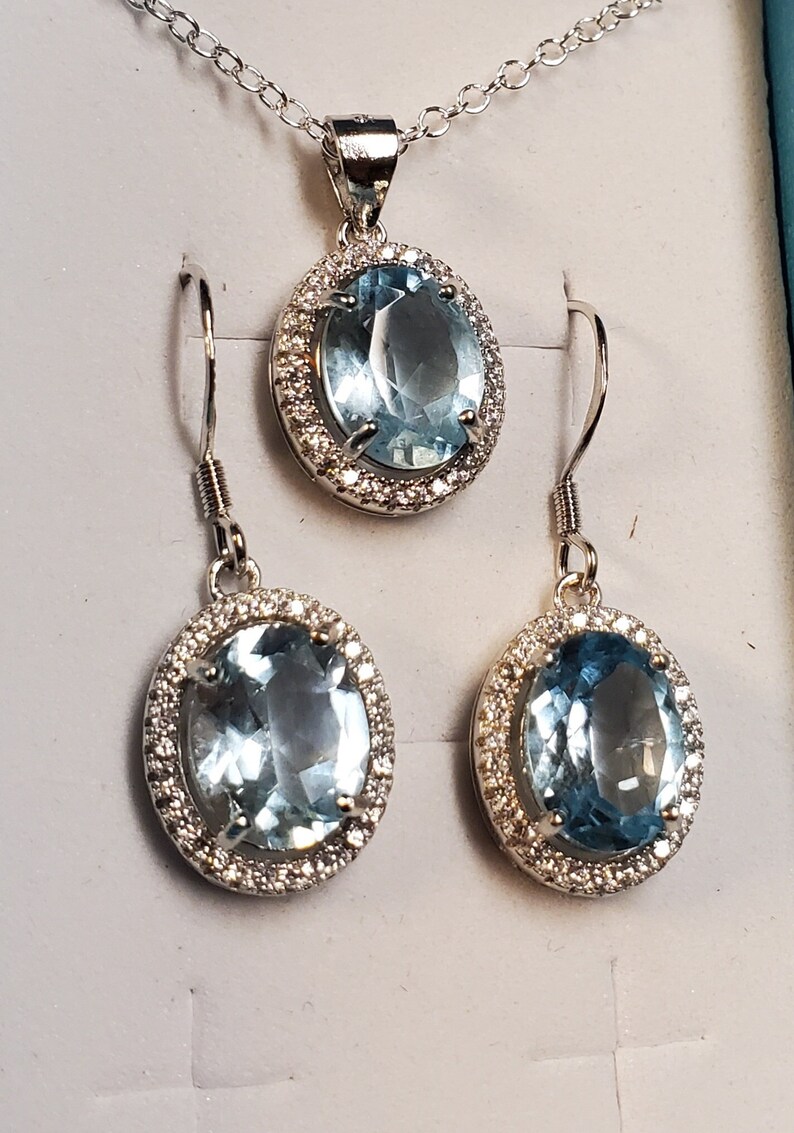 May include: A silver-toned jewelry set featuring a pendant necklace and matching earrings. Each piece showcases a large, oval, light blue gemstone surrounded by a halo of small, clear stones. The set is displayed on a light gray surface.