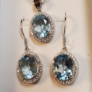 May include: A jewelry set featuring a pendant necklace and a pair of earrings. Each piece has a large, oval, light blue gemstone surrounded by small, clear stones. The metal appears to be silver-colored.