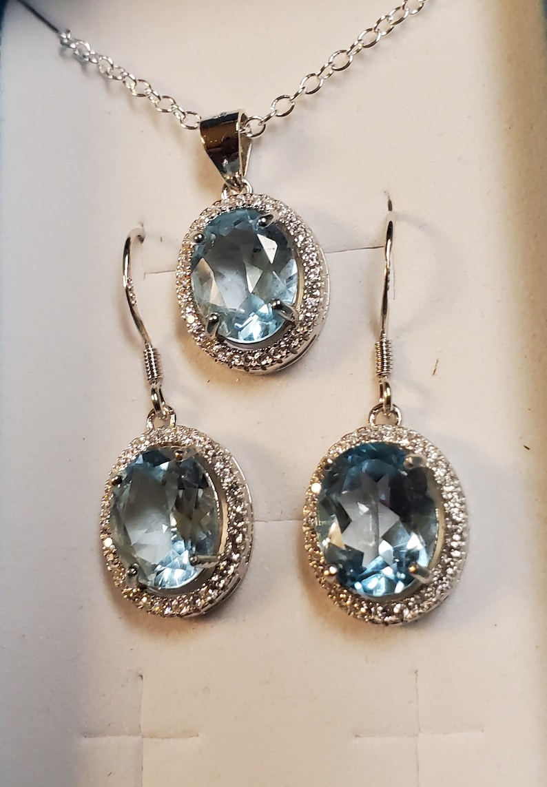 May include: A jewelry set featuring a pendant necklace and a pair of earrings. Each piece has a large, oval, light blue gemstone surrounded by small, clear stones. The metal appears to be silver-colored.