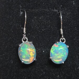 Pu&ograve; includere: Un paio di orecchini pendenti con opali di forma ovale incastonati in griffe di metallo color argento. Ogni opale mostra un vivace gioco di colori con riflessi verdi, blu e arancioni. Gli orecchini pendono da semplici ganci in metallo.