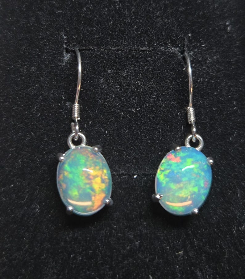 Pu&ograve; includere: Un paio di orecchini pendenti con opali di forma ovale incastonati in griffe di metallo color argento. Ogni opale mostra un vivace gioco di colori con riflessi verdi, blu e arancioni. Gli orecchini pendono da semplici ganci in metallo.