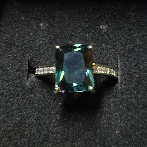 May include: A silver ring with a large, emerald-cut, dark blue gemstone surrounded by smaller, clear gemstones.