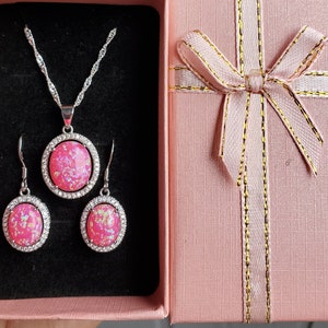 Puede incluir: Un conjunto de collar y pendientes de &oacute;palo rosa en un engaste plateado. Las joyas se muestran en una caja de regalo rosa con una cinta dorada.