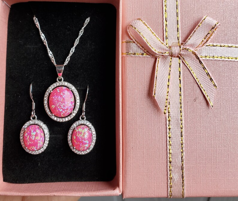 Puede incluir: Un conjunto de collar y pendientes de &oacute;palo rosa en un engaste plateado. Las joyas se muestran en una caja de regalo rosa con una cinta dorada.