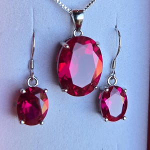 May include: A jewelry set consisting of a silver necklace with an oval-shaped, faceted red gemstone pendant and a pair of matching oval-shaped, faceted red gemstone drop earrings with silver hooks, all displayed against a plain white background.
