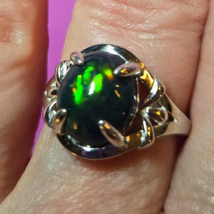 May include: A silver ring featuring a large, oval-shaped black opal gemstone with vibrant green and yellow flashes. The opal is set in a decorative silver band with a twisted design and claw-like prongs.