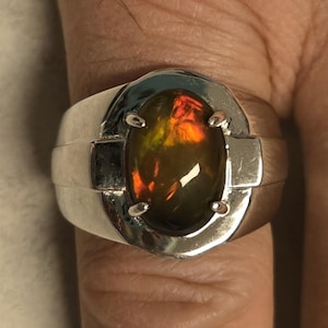 May include: A silver-toned ring featuring a large, oval-shaped dark stone with vibrant orange and green iridescent flashes. The stone is held in a four-prong setting within a wide, polished metal band that tapers slightly toward the sides.