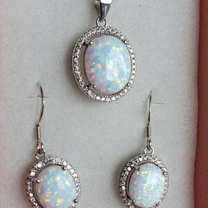 May include: A silver necklace and earrings set with a white opal stone in each piece. The stones are surrounded by a halo of smaller clear stones.