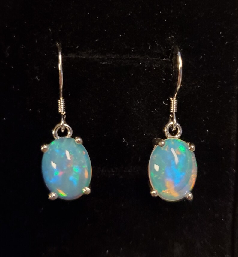 Pu&ograve; includere: Un paio di orecchini pendenti con opali ovali e iridescenti incastonati in griffe di metallo color argento. Ogni orecchino pende da un semplice gancio color argento, mostrando una base bianco latte con bagliori di luce blu, verde e arancione.