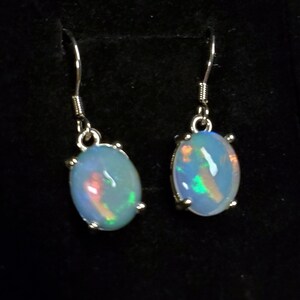 Pu&ograve; includere: Un paio di orecchini pendenti con opali ovali bianco latte con bagliori iridescenti verdi e arancioni. Ogni pietra &egrave; montata in una montatura a griffe in metallo color argento e pende da un semplice gancio per orecchie in metallo color argento.