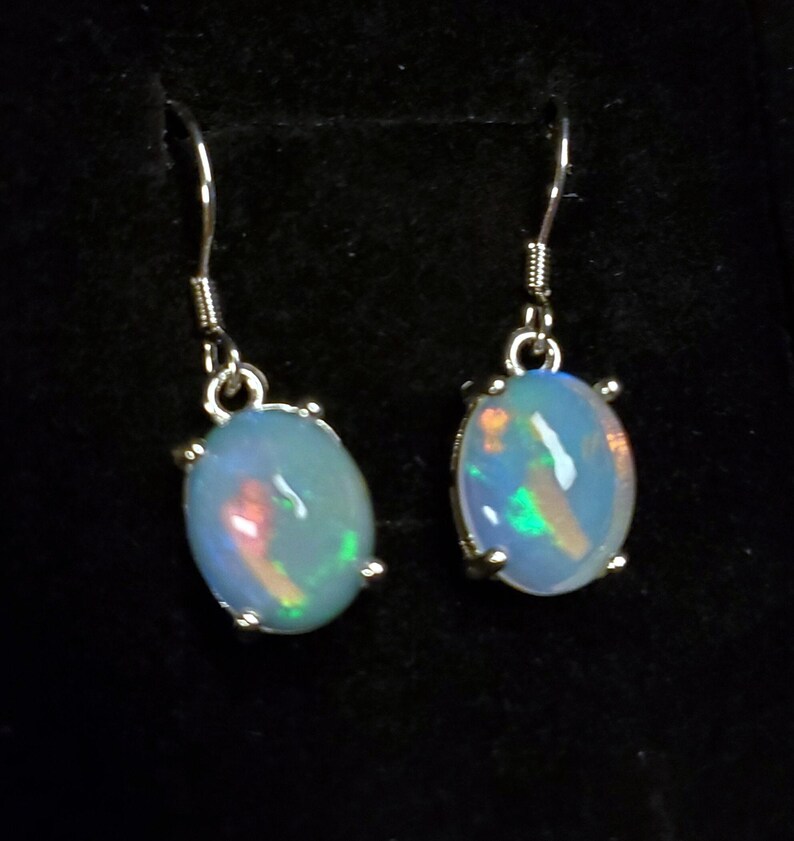 Pu&ograve; includere: Un paio di orecchini pendenti con opali ovali bianco latte con bagliori iridescenti verdi e arancioni. Ogni pietra &egrave; montata in una montatura a griffe in metallo color argento e pende da un semplice gancio per orecchie in metallo color argento.
