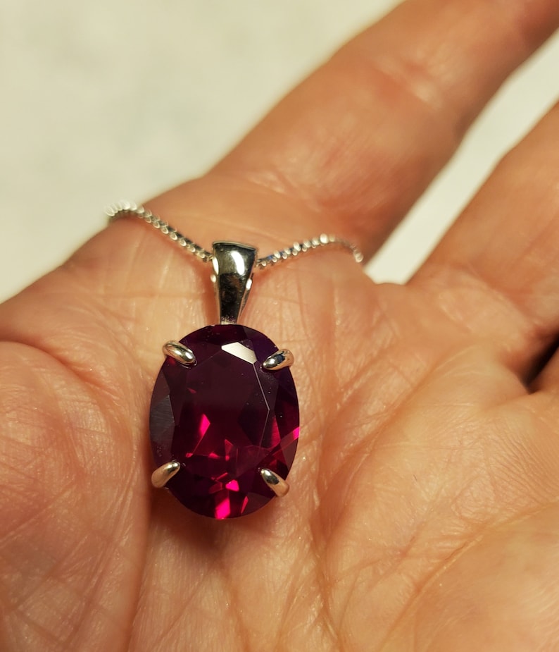 Puede incluir: Un collar de cadena de plata con un colgante de rub&iacute; de forma ovalada. El rub&iacute; es de color rojo intenso y tiene un corte facetado.
