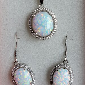 May include: A silver necklace and earrings set with a white opal pendant and matching earrings. The opal is surrounded by a halo of small clear stones.