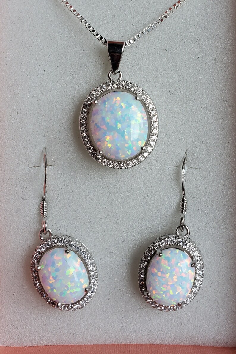 May include: A silver necklace and earrings set with a white opal pendant and matching earrings. The opal is surrounded by a halo of small clear stones.