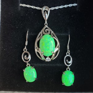 May include: A silver necklace and earrings set with a green opal pendant and matching earrings. The pendant features a delicate, ornate design with a swirl pattern.