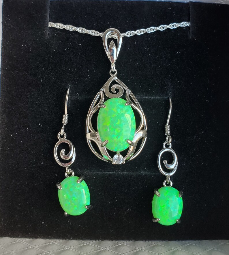May include: A silver necklace and earrings set with a green opal pendant and matching earrings. The pendant features a delicate, ornate design with a swirl pattern.