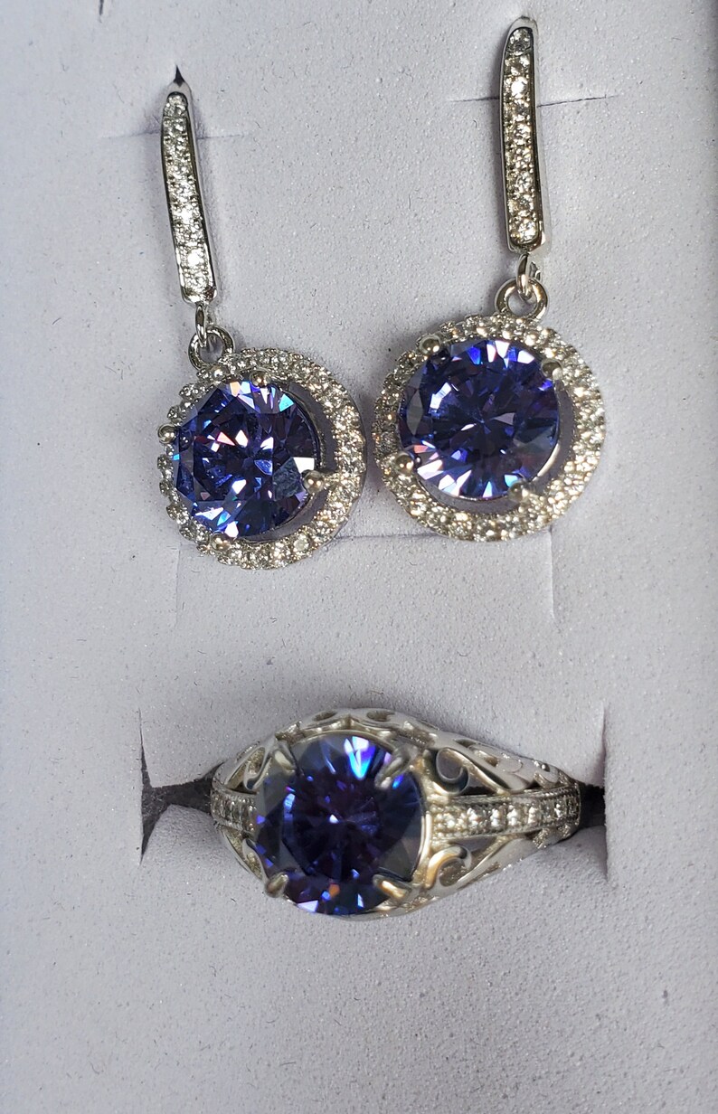 May include: A silver ring and a pair of earrings featuring a large, round, purple gemstone encircled by smaller, clear gemstones.