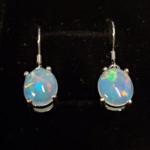 Pu&ograve; includere: Un paio di orecchini pendenti con opali cabochon di forma ovale incastonati in griffe di metallo color argento. Gli opali presentano un colore di base bianco latte con riflessi iridescenti di luce blu, verde e arancione.