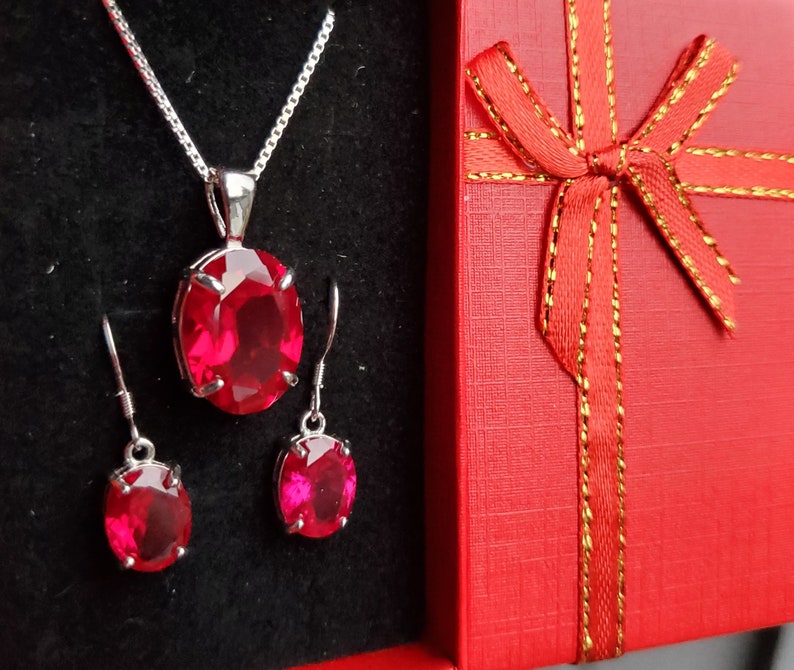 Puede incluir: Un conjunto de joyas de piedras preciosas rojas con un collar colgante y pendientes. El conjunto est&aacute; empaquetado en una caja de regalo roja con una cinta dorada.