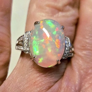 May include: An oval-shaped opal ring with a silver band. The opal has a milky white base with flashes of green, orange, and yellow. The band is adorned with small, clear gemstones on each side of the opal.