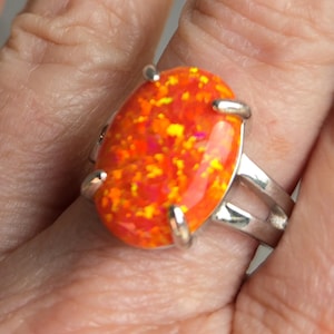 May include: A silver ring featuring a large, oval-shaped opal. The opal has a vibrant, fiery color palette of red, orange, and yellow hues. The ring's band splits into two parallel lines on the sides of the opal.
