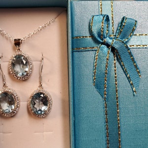 May include: A silver necklace and earring set with oval, light blue gemstones and a halo of small, clear stones. The jewelry is presented in a light blue gift box with a matching ribbon and gold trim.