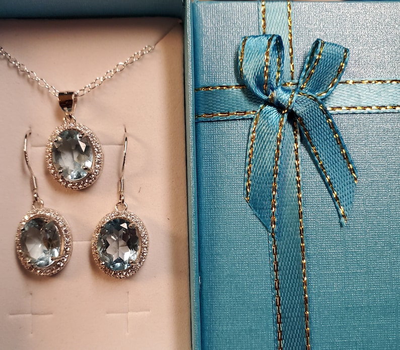 May include: A silver necklace and earring set with oval, light blue gemstones and a halo of small, clear stones. The jewelry is presented in a light blue gift box with a matching ribbon and gold trim.