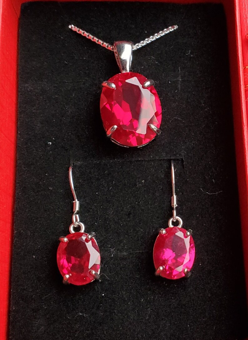 Puede incluir: Un collar de plata con un colgante de piedra roja ovalada grande y pendientes a juego. Las joyas se muestran en una caja de terciopelo rojo.