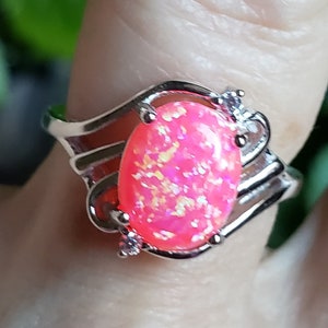 May include: A silver ring with a pink oval-shaped opal gemstone and small clear accents. The ring has a delicate, intricate design.