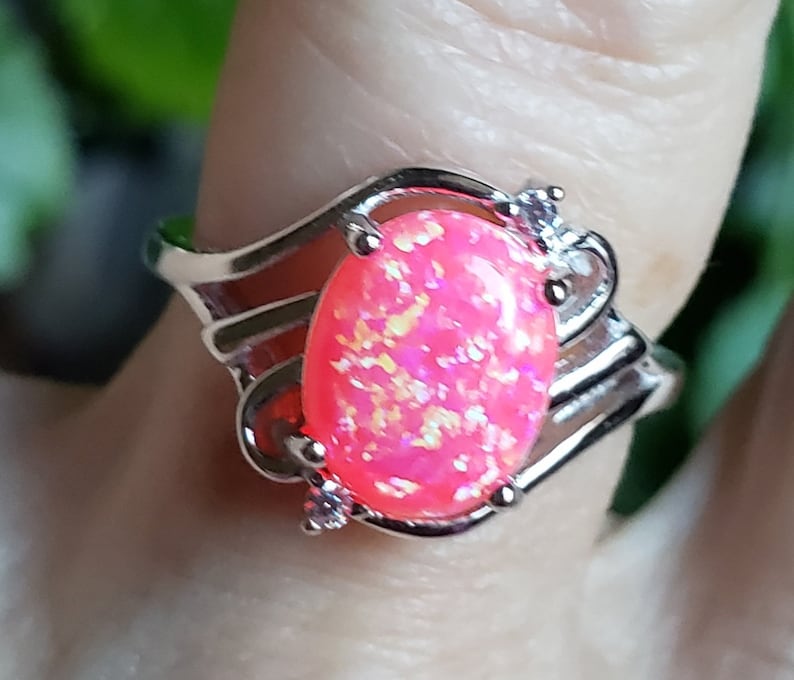 May include: A silver ring with a pink oval-shaped opal gemstone and small clear accents. The ring has a delicate, intricate design.
