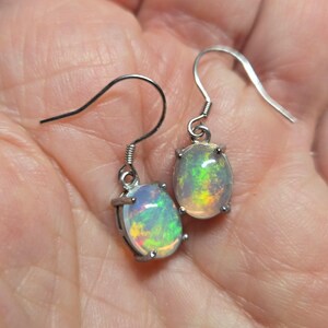 Pu&ograve; includere: Un paio di orecchini pendenti color argento con opali ovali. Ogni opale presenta una base bianca traslucida con vivaci bagliori di fuoco verde, giallo e arancione. Le pietre sono fissate con quattro griffe a ganci auricolari curvi.