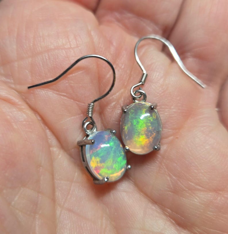 Pu&ograve; includere: Un paio di orecchini pendenti color argento con opali ovali. Ogni opale presenta una base bianca traslucida con vivaci bagliori di fuoco verde, giallo e arancione. Le pietre sono fissate con quattro griffe a ganci auricolari curvi.
