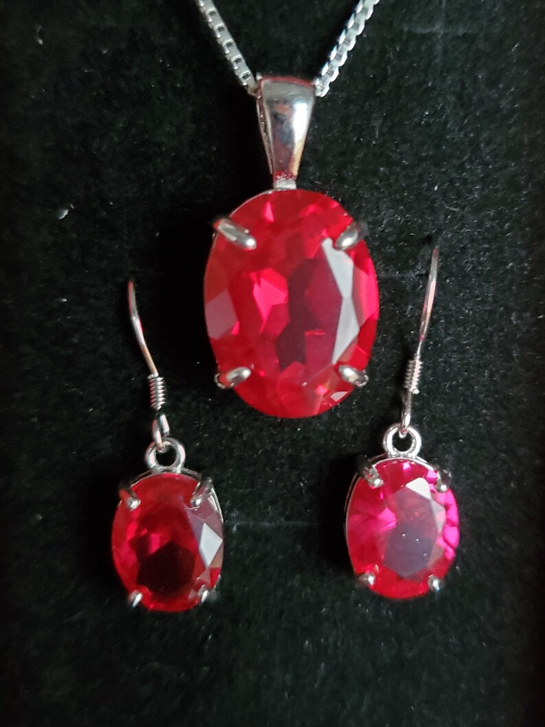 Puede incluir: Un collar de plata con un colgante ovalado rojo y pendientes a juego. El colgante y los pendientes presentan una gran piedra roja facetada.