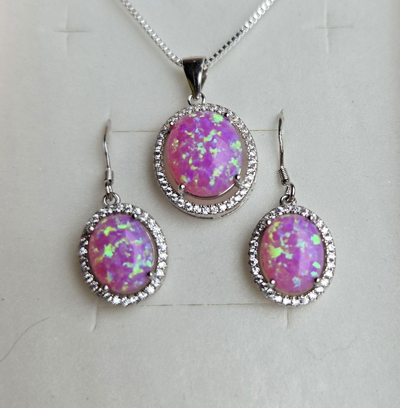 May include: A pink opal necklace and earrings set with a silver chain and oval-shaped stones surrounded by a halo of clear cubic zirconia.