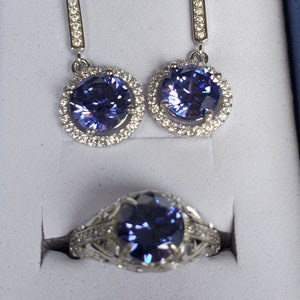 May include: A set of amethyst jewellery featuring a ring and a pair of earrings. The ring and earrings have a silver setting with a round, purple amethyst stone surrounded by smaller clear stones.