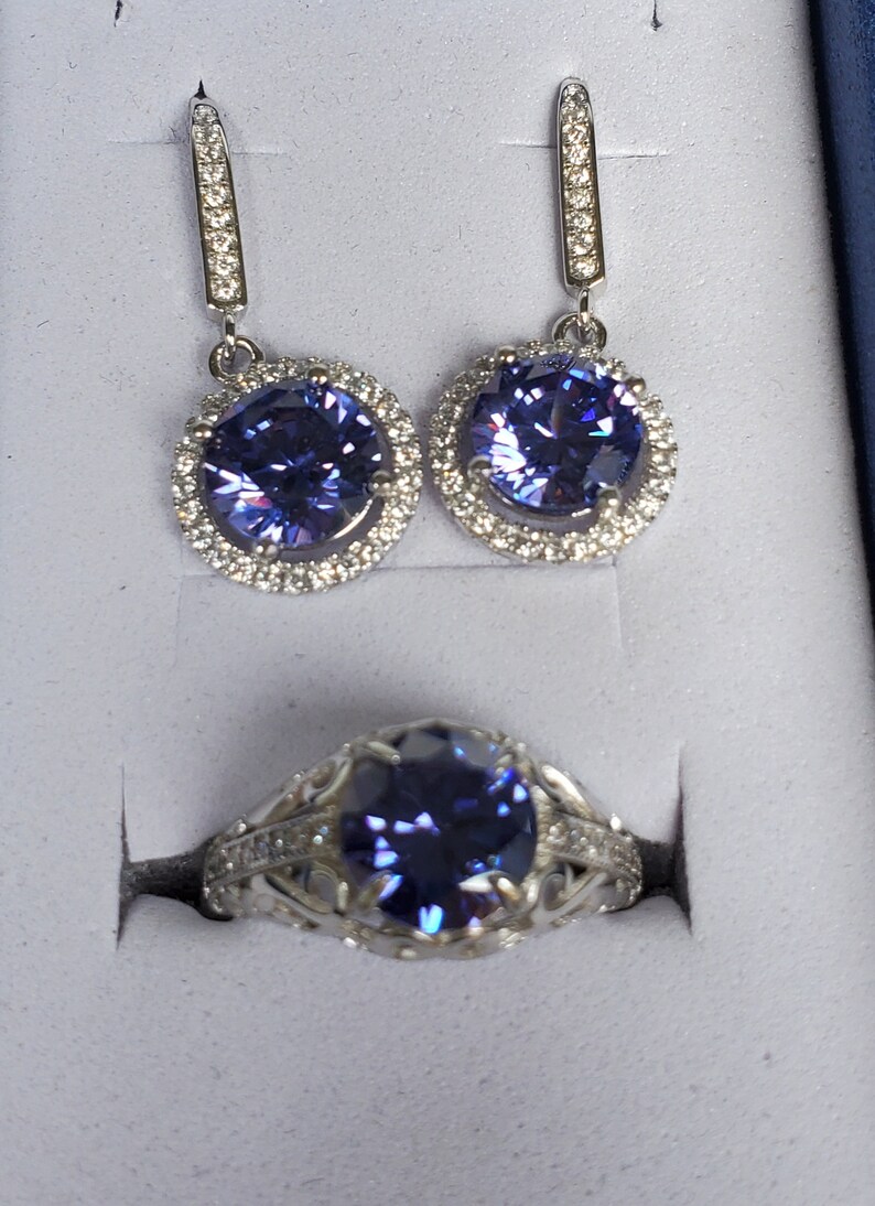 May include: A set of amethyst jewellery featuring a ring and a pair of earrings. The ring and earrings have a silver setting with a round, purple amethyst stone surrounded by smaller clear stones.