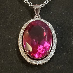 May include: A silver chain necklace with a pendant featuring a large oval-shaped pink gemstone surrounded by a halo of smaller clear gemstones.